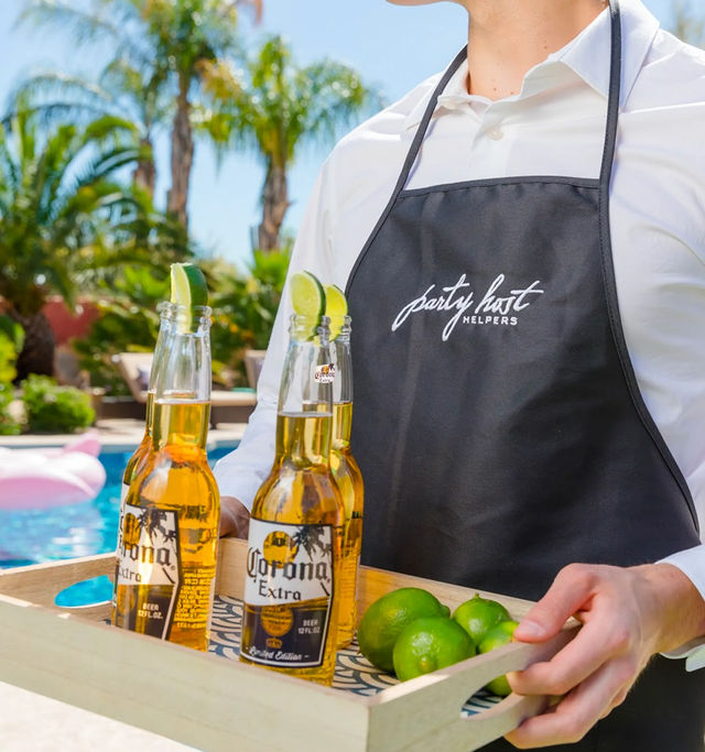 Sunny poolside scene: server in a black apron carrying a wooden tray with three lime-garnished chilled lager bottles and fresh limes, palm trees and backyard pool in the background.