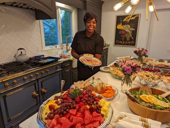 Smiling server in a modern home kitchen holding a platter of crackers and dip beside a kitchen island piled with a fruit platter, salads, charcuterie and floral centerpieces for a buffet spread.