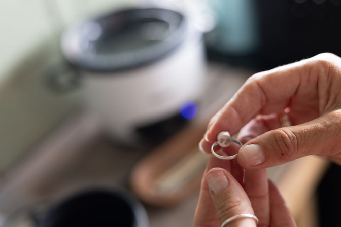Close-up of hands inspecting a silver ring with a small pearl over a blurred home workbench, jewelry repair/inspection scene