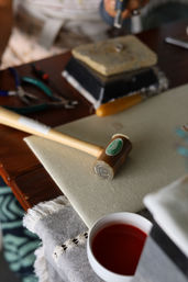 Close-up of a craft workbench: wooden mallet, green-handled pliers, bench block and tools on a fabric-covered table next to a cup of red tea.