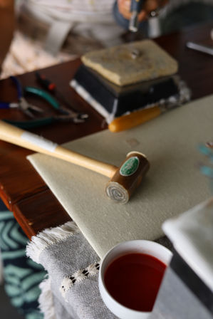 Close-up of a wooden mallet on canvas at a craft workbench, with pliers, a soldering block and a small white cup of red liquid nearby.