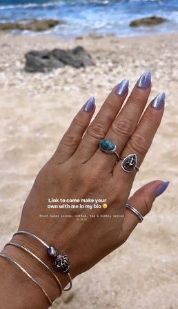 Close-up of a hand posed against a sandy beach and blue ocean, showing lavender shimmery almond nails, silver rings and shell bracelet with white overlay text about a link and refreshments.