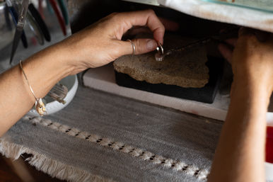 Close-up of hands shaping a silver ring on a soldering stone at a jewelry workbench, with tools and a woven cloth mat nearby