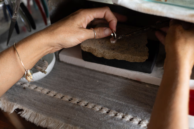 Close-up of hands shaping a silver ring on a soldering stone at a jewelry workbench, with tools and a woven cloth mat nearby