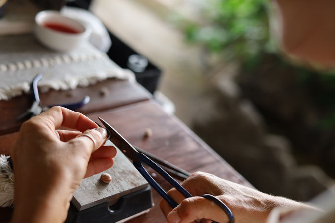 Close-up of hands using blue-handled pliers to shape a small bead on a wooden workbench, jewelry-making tools, a cup, and blurred greenery in the background.