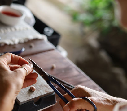 Close-up of hands using blue-handled pliers to shape a small bead on a wooden workbench, jewelry-making tools, a cup, and blurred greenery in the background.