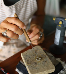 Close-up of a jeweler's hands shaping a silver ring with a wooden-handled pick on a firebrick soldering block beside a bench torch in a jewelry workshop.