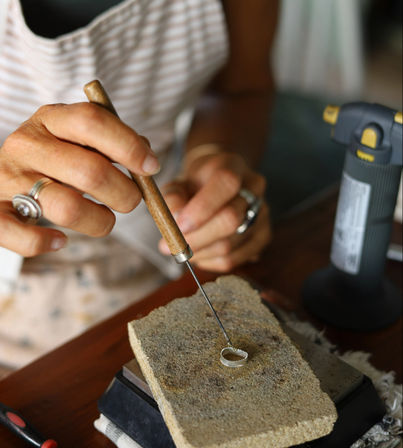 Close-up of a jeweler's hands shaping a silver ring with a wooden-handled pick on a firebrick soldering block beside a bench torch in a jewelry workshop.