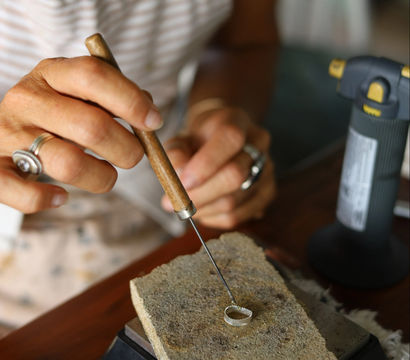 Close-up of a jeweler's hands shaping a silver ring with a wooden-handled pick on a firebrick soldering block beside a bench torch in a jewelry workshop.