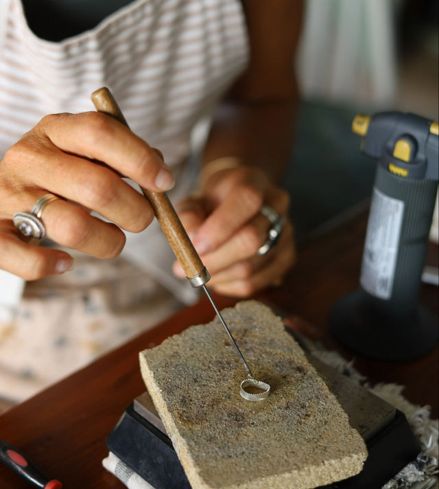 Close-up of a jeweler's hands shaping a silver ring with a wooden-handled pick on a firebrick soldering block beside a bench torch in a jewelry workshop.