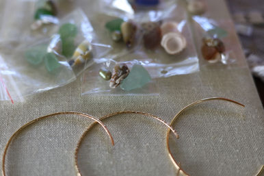 Close-up of coastal-inspired jewelry components: sea glass and tiny seashell beads in clear bags with three gold hoop earring frames on a linen craft surface