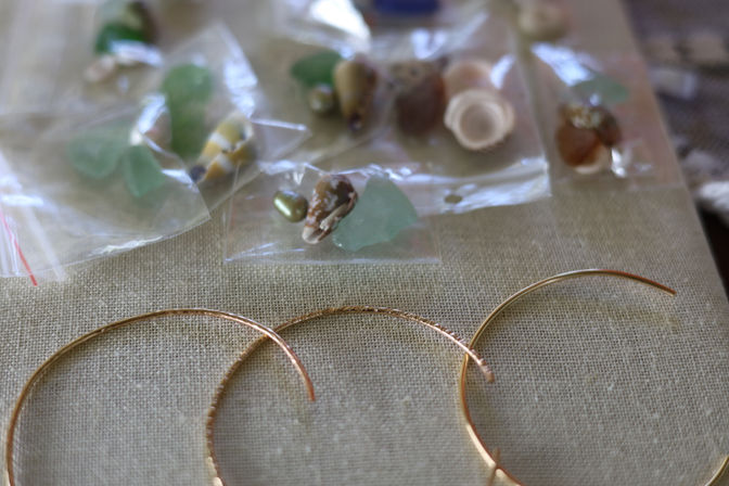 Close-up of coastal-inspired jewelry components: sea glass and tiny seashell beads in clear bags with three gold hoop earring frames on a linen craft surface