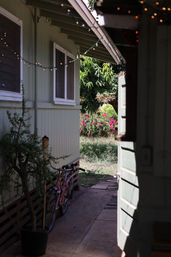 Narrow backyard pathway between a pale-green wooden house and open door, with a pink bicycle, potted plant, string lights, and a sunlit garden of pink bougainvillea and grass beyond.