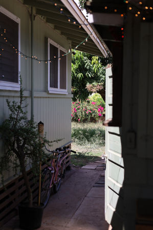 Narrow backyard pathway between a pale-green wooden house and open door, with a pink bicycle, potted plant, string lights, and a sunlit garden of pink bougainvillea and grass beyond.
