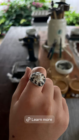Close-up of a hand wearing a spotted shell ring set in silver, blurred artisan workspace in the background with brushes, jars and greenery — handmade shell ring on a studio worktable.