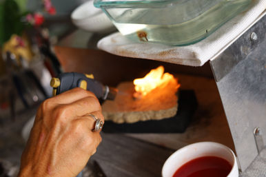 Close-up of a hand wearing a silver ring using a butane torch to heat a firebrick, bright orange flame on an artisan workbench with a glass dish and a cup of red liquid nearby.