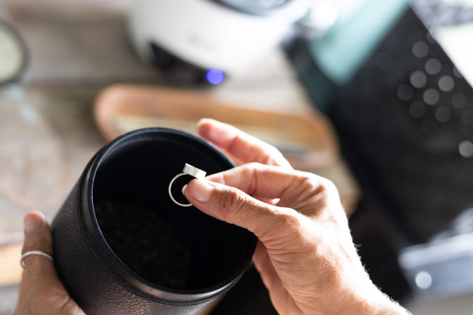 Close-up of hands placing a silver ring into a black cylindrical container on a blurred desk, a casual jewelry storage moment.
