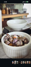 Close-up of a white ramekin filled with mixed purple, brown and spotted seashells on a woven placemat, with a blurred white bowl and kitchen table in the background — beachy tabletop decor.