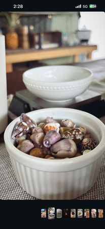 Close-up of a white ramekin filled with mixed purple, brown and spotted seashells on a woven placemat, with a blurred white bowl and kitchen table in the background — beachy tabletop decor.