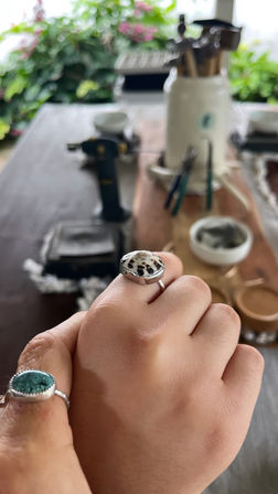 Close-up of a hand wearing two handmade silver rings — a speckled white cabochon and a green-blue stone — held over a wooden jewelry workbench with pliers, a torch and blurred plants in the background.