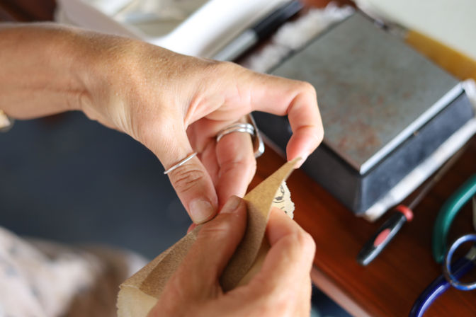 Close-up of hands wearing silver rings hand-sanding a small wooden piece with sandpaper at a DIY workbench with tools nearby.