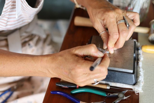 Close-up of a jeweler’s hands filing a silver ring on a metal bench block, surrounded by pliers, cutters and other small crafting tools on a wooden workbench