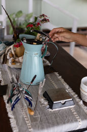 Hand reaching for pliers in a turquoise enamel pitcher repurposed as a tool caddy, surrounded by jewelry-making pliers, a small vase of pink flowers and seashells on a linen table runner