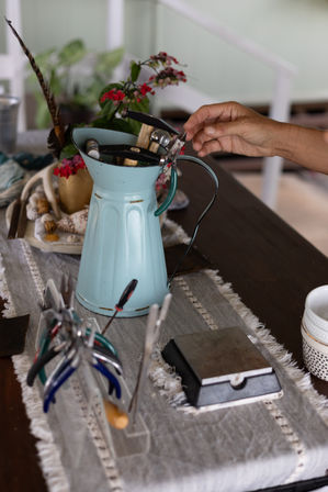 Hand reaching for pliers in a vintage turquoise pitcher repurposed as a tool holder on a home craft table, surrounded by jewelry pliers, files, a bench block and small pink flowers.