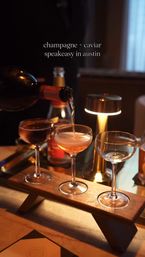 Three coupe glasses on a wooden tasting board with champagne being poured into the center glass at a cozy, lamp-lit Austin speakeasy — elegant champagne flight vibe.