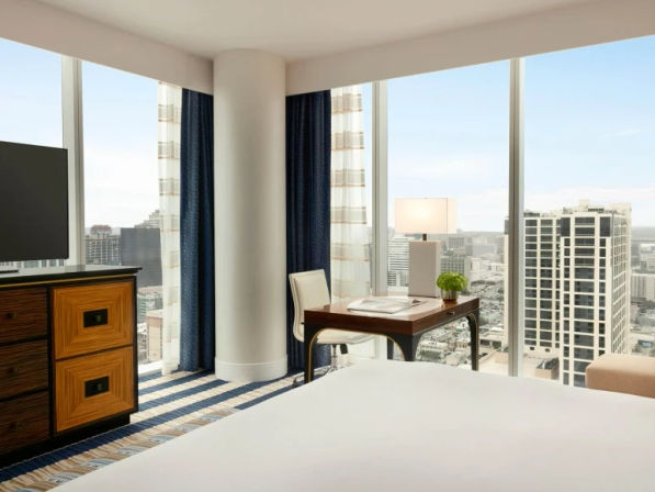 Bright modern hotel room with floor-to-ceiling windows overlooking downtown high-rise skyline, king bed in foreground, desk with lamp and chair, and TV on a wooden dresser