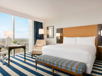Bright modern hotel room with king bed, striped blue rug, bench, desk and expansive city skyline view