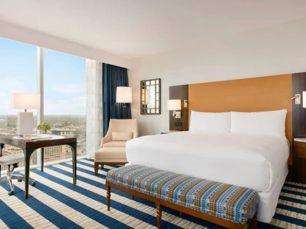 Bright modern hotel room with king bed, striped blue rug, bench, desk and expansive city skyline view