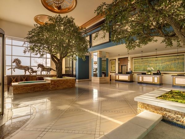 Sunlit, spacious luxury hotel lobby with large indoor trees, gleaming marble floors, galloping horse sculptures and reception desks