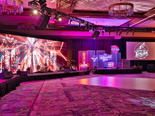 Hotel ballroom event stage bathed in pink and purple light with a large LED backdrop, band instruments and drum kit, neon club-style Texas-themed signage, and a central dance floor.