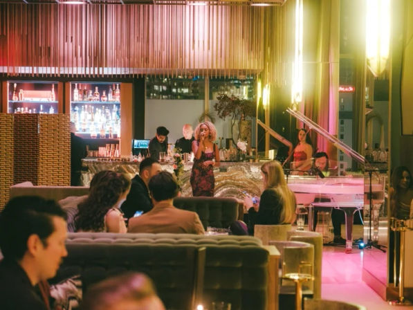 Chic city lounge scene with a live singer and pianist performing by a glowing marble bar, patrons on velvet sofas sipping cocktails under warm pink-and-gold lighting and backlit liquor shelves.