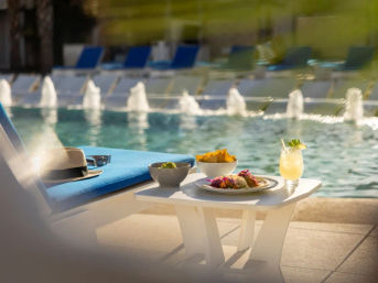 Sunlit poolside scene with a white table holding tacos, chips, guacamole and a lime cocktail beside a blue lounge chair topped with a straw hat and sunglasses, fountain jets in the background.