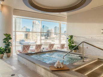 Sunny indoor luxury spa hot tub with bubbling jacuzzi, floor-to-ceiling windows framing a downtown skyline, pink lounge chairs, potted plants and stacked rolled towels.