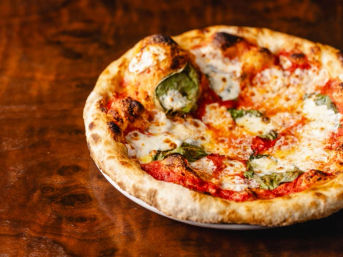 Neapolitan-style margherita pizza with charred, puffy wood-fired crust, bubbling mozzarella, bright tomato sauce and fresh basil on a white plate atop a rustic wooden table.