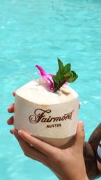 Hands holding a fresh coconut drink topped with a pink orchid and mint beside a turquoise pool in Austin, Texas.