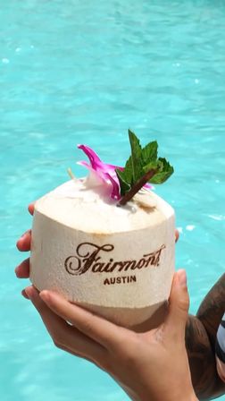 Hands holding a fresh coconut drink topped with a pink orchid and mint beside a turquoise pool in Austin, Texas.