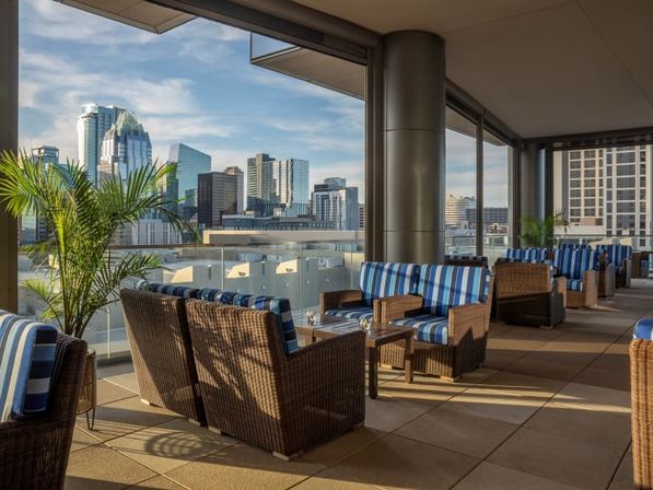 Sunlit rooftop lounge with wicker sofas and blue-striped cushions, potted palms and tables overlooking a downtown high-rise skyline.