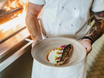 Chef in a restaurant kitchen holding a ceramic plate with a seared fish fillet (charred skin), white cream and orange puree, garnished with pink edible flowers and yellow microgreens, wood-fired oven flames visible in the background.