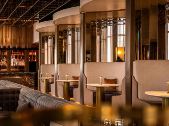 Luxury hotel bar lounge with curved private booths, brass-accented round tables, tufted velvet sofa and backlit liquor shelves glowing in warm ambient lighting — stylish, inviting cocktail setting.