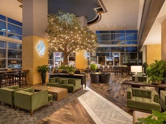 Modern hotel lobby in an urban downtown setting with a large indoor tree wrapped in string lights, green velvet sofas and cozy seating areas, wooden herringbone floor, bar and floor-to-ceiling windows showing city skyline at night.