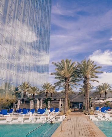 Rooftop resort-style pool with white lounge chairs and blue cushions, palm trees along a wooden deck beside a reflective glass skyscraper under a bright blue sky