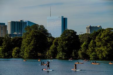 Paddleboarders and kayakers on a calm urban lake, lush green shoreline in the foreground and glass skyscrapers forming a downtown skyline under a blue sky.