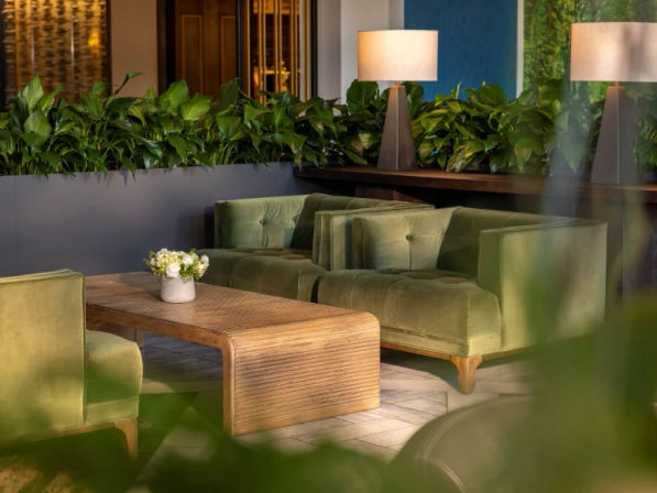 Cozy green-velvet lounge seating in a hotel lobby: tufted sofas, curved wooden coffee table with white flower arrangement, leafy indoor plants and warm table lamps.