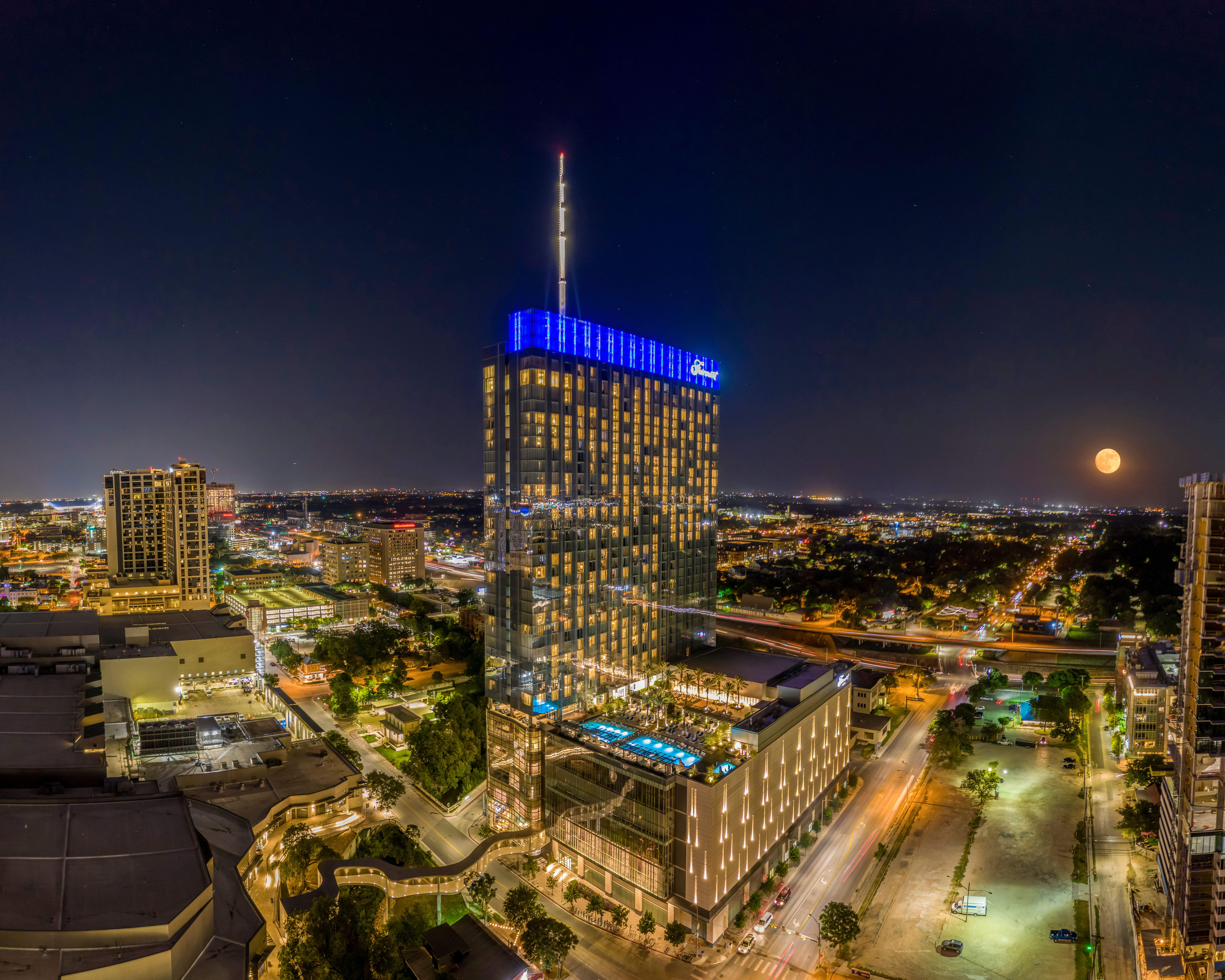Nighttime urban skyline featuring a modern glass high-rise with blue crown lights and rooftop pool, city streets aglow and a full moon on the horizon.