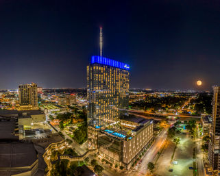 Nighttime urban skyline featuring a modern glass high-rise with blue crown lights and rooftop pool, city streets aglow and a full moon on the horizon.