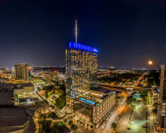 Nighttime urban skyline featuring a modern glass high-rise with blue crown lights and rooftop pool, city streets aglow and a full moon on the horizon.